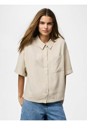 Kobieta o długich brązowych włosach, ubrana w beżową koszulę z krótkim rękawem na guziki i niebieskie dżinsy, stojąca na jednolitym białym tle.