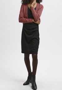 Kvinna klädd i en svart klänning med draperad design, kombinerad med en rosa kcardigan och svarta ankelskor, poserar mot en enfärgad bakgrund.