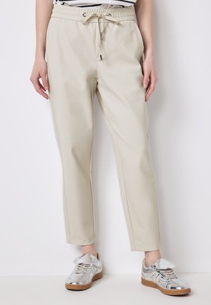 Beige jogger pants dengan pinggang elastis, penutupan tali, saku samping, potongan menyempit, tekstur halus, dipadukan dengan sepatu sneaker perak.