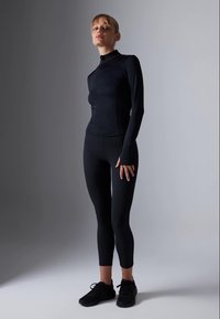 Žena stoji u crnoj uskoj sportskoj odjeći i crnim tenisicama naspram sive gradijentne pozadine.