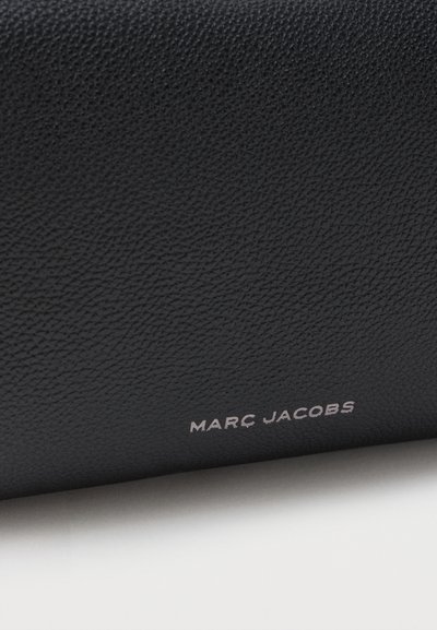 Μαύρη υφασμάτινη επιφάνεια δέρματος με μικρό ασημένιο κείμενο "MARC JACOBS" κοντά στο κάτω άκρο.