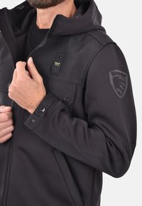 Zwarte softshell jas met een ritssluiting aan de voorkant, drukknopen en een gestructureerd schouderlabel. Voorzien van een capuchon en praktische zakken.