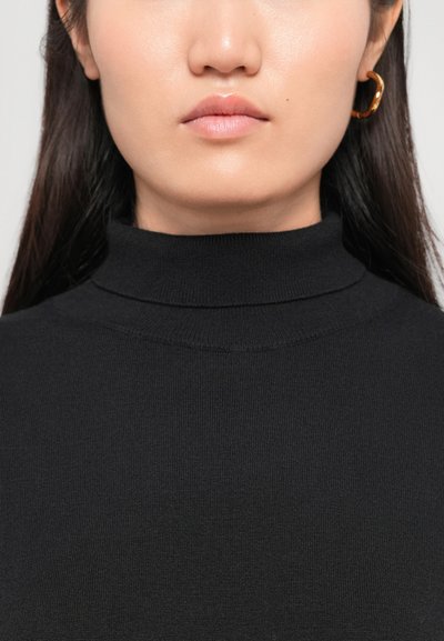 Schwarzer gerippter Rollkragenpullover mit taillierter Passform. Das Modell trägt goldene Creolen. Die glatte Hauttextur und die neutrale Lippenfarbe sind sichtbar.