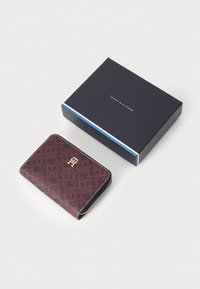Tommy Hilfiger wallet in a maroon patterned material with a gold logo and zipper, next to a dark blue box with logo details.
