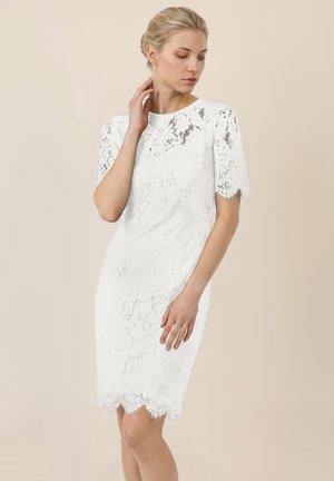 Vrouw in een witte kanten jurk tot op de knie met korte mouwen, lichte beige achtergrond, kijkt naar beneden, hand bij hals en heup.