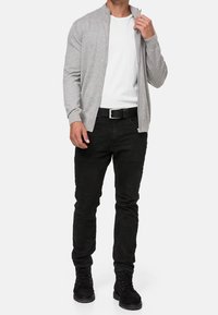 Šedý cardigan na zip přes bílé tričko, doplněný černými džínami a černými botami. Textura je hladká, s přiléhavým střihem a žebrovaným límcem.