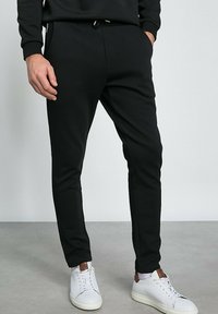 Des joggers noirs fuselés en tissu doux présentent une taille à cordon de serrage et des poches zippées, associés à des baskets blanches.