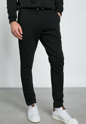 Des joggers noirs fuselés en tissu doux présentent une taille à cordon de serrage et des poches zippées, associés à des baskets blanches.