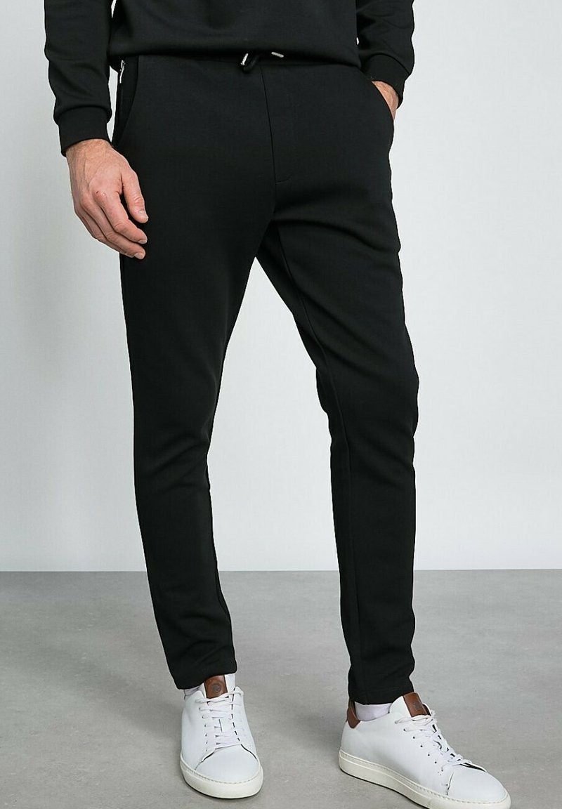 Des joggers noirs fuselés en tissu doux présentent une taille à cordon de serrage et des poches zippées, associés à des baskets blanches.