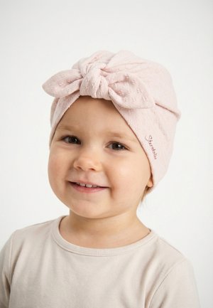 Kleinkind lächelt, trägt ein hellrosa strukturiertes Turban-Stirnband mit Schleife und ein blassbeiges Hemd vor weißem Hintergrund.