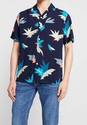Hombre vistiendo una camisa azul marino de manga corta abotonada con patrones abstractos de hojas multicolores y vaqueros azules.