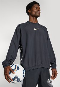 Svart langermet sportstopp med rund hals, elastiske mansjetter og hvite detaljer; person holder en mønstrete fotball med blå og oransje detaljer.