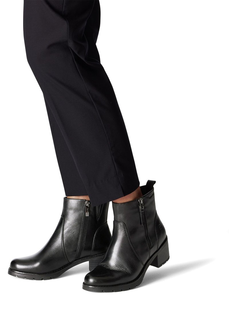 Persona que lleva botas de tobillo negras con cremalleras laterales y tacones bajos, combinadas con pantalones ajustados negros.