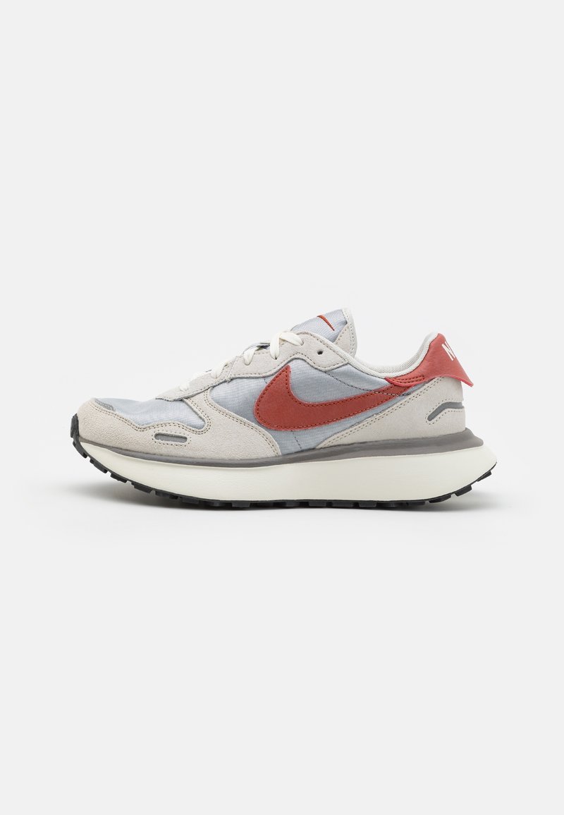 Baskets Nike blanches et gris clair avec logo swoosh rouge, accents en suède beige et semelle blanche, présentées sur un fond clair uni.