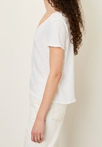 Kvinna med långt, lockigt hår som bär en vit kortärmad topp och ljusa byxor, sedd från sidan mot en enfärgad bakgrund.