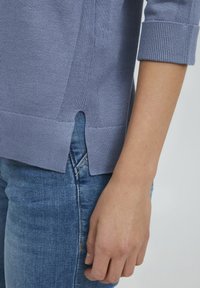 Hellblaues Strickpullover mit Umschlagärmel, seitlichen Schlitzen, geripptem Saum, getragen über Jeans und zeigt eine weiche Textur und einen lässigen Stil.