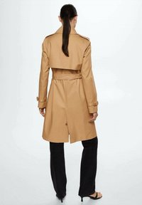 Beżowy trencz o skróconym kroju, z pagonami na ramionach, paskiem w talii i wentylowanym tyłem, zestawiony z czarnymi spodniami i sandałami.