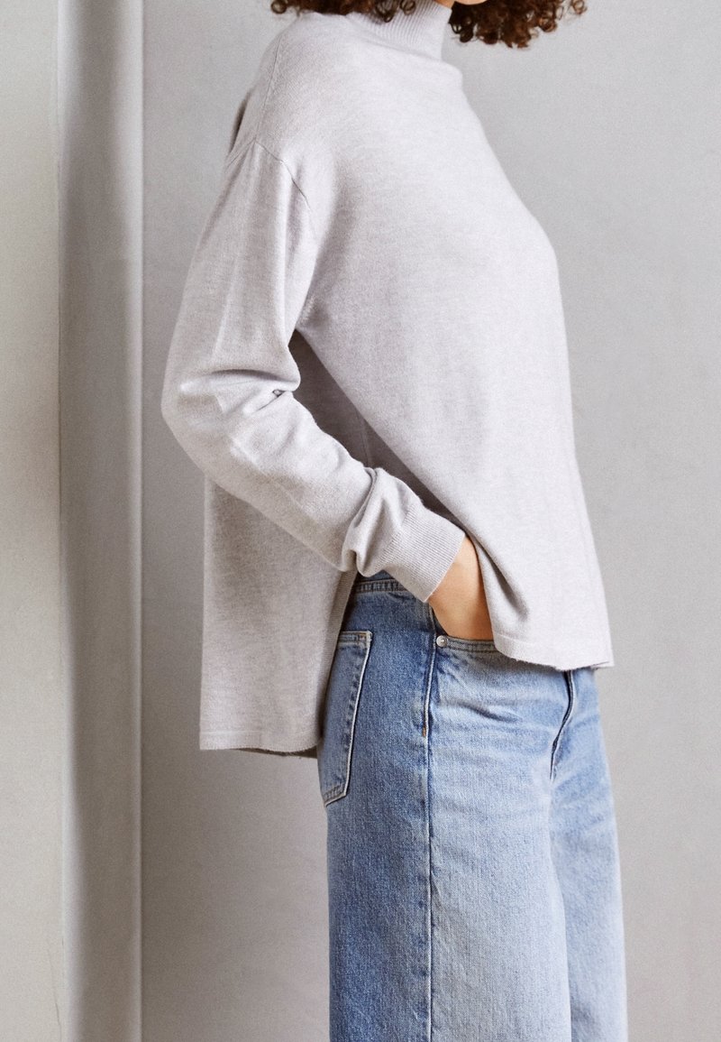 Personne aux cheveux bouclés portant un pull à col roulé gris clair et un jean bleu, une main dans la poche, sur un fond neutre.
