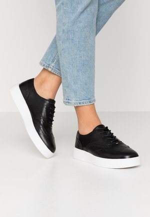Pieds portant des baskets montantes en cuir noir avec semelles blanches et des jeans bleu clair retroussés sur un sol blanc uni.