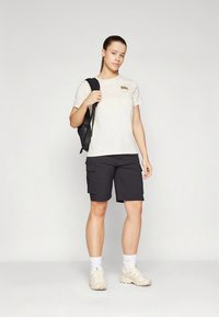 Ljusbeige t-shirt med en liten logotyp, svarta cargoshorts och cremefärgade sneakers, bärs med en svart ryggsäck. Enkel, funktionell utomhusoutfit.