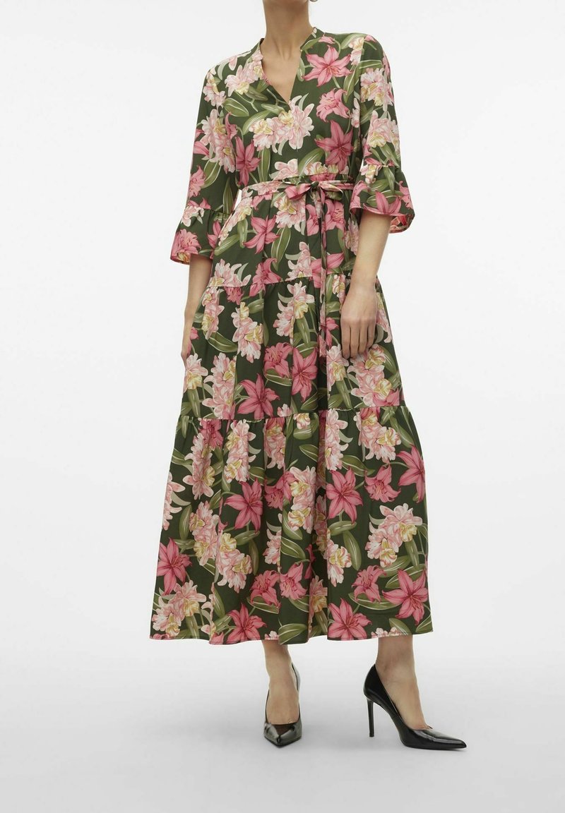 Femme portant une robe portefeuille verte à fleurs roses et crème, manches trois-quarts, et chaussures à talons noires.