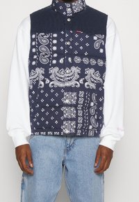 Mörkblå väst med vita paisley- och blommönster, manchesterkrage, knappstängning, bärs över en vit långärmad skjorta.