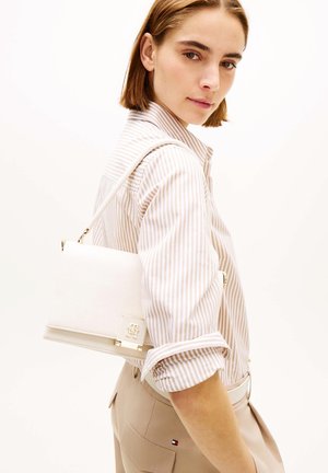 Frau trägt beige gestreiftes Hemd und Hose, trägt eine weiße Ledertasche mit goldener Verschluss auf der Schulter.