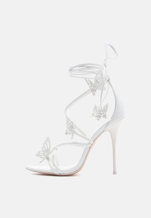Λευκό σανδάλι ψηλού τακουνιού με λεπτά λουράκια και διακοσμητικές τρύπες σε σχήμα πεταλούδας, με κλείσιμο δετό γύρω από τον αστράγαλο.