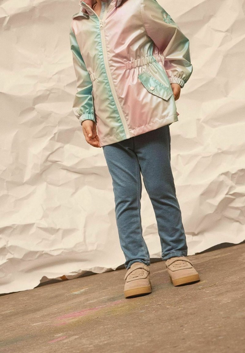 Veste multicolore irisée avec une taille élastique, associée à des leggings bleus et des chaussures beiges. Tissu texturé avec capuche et fermeture éclair.