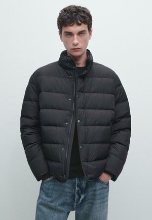 Schwarze Puffjacke aus gepolstertem Stoff mit Druckknopfverschluss und hohem Kragen. Trägt eine schwarze Bluse und ist mit blauen Jeans kombiniert.