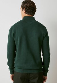 Pulover verde tricotat cu guler înalt și manșete și tiv ondulat. Materialul pare texturat, oferind o potrivire strânsă.