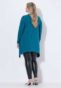 Miętowozielona bluzka z długim rękawem i asymetrycznym dołem, zestawiona z czarnymi skórzanymi legginsami i srebrnymi sandałami na obcasie. Gładka faktura, luźny krój.