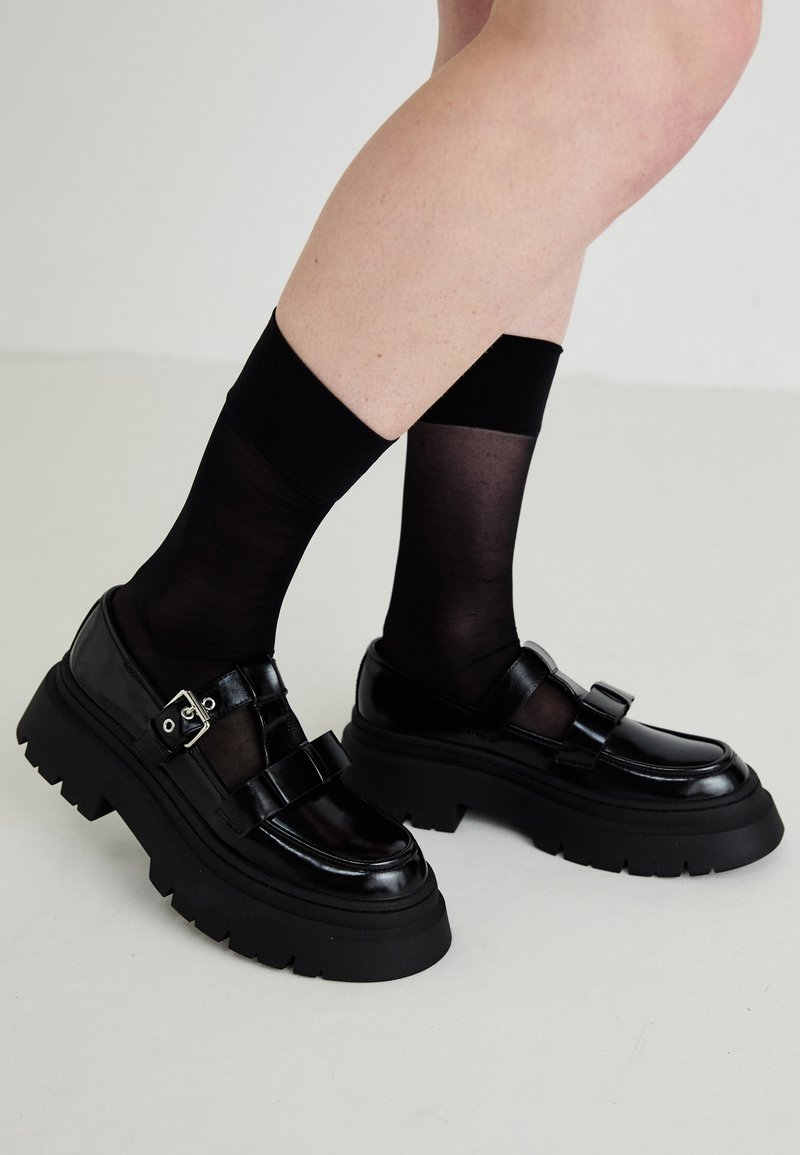 Zapatos de charol negro con suelas gruesas, dos correas decorativas y una hebilla, combinados con calcetas negras transparentes.