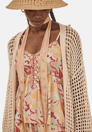 Cárdigan de ganchillo beige claro sobre un vestido de colores con estampados florales, adornado con botones y una correa trenzada en el cuello.