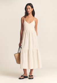 Linen blend maxi dress in cream with a V-neckline, adjustable spaghetti straps, gathered waist, and tiered hem. Paired with a woven tote and sandals.