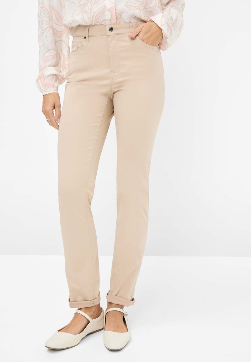 Pantalon beige slim-fit avec une texture lisse, poches avant et ourlet retroussé, associé à des ballerines blanches.