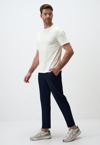 Homme portant un t-shirt blanc uni à manches courtes, un pantalon bleu marine et des baskets beiges, se tenant avec une main dans sa poche devant un fond blanc.