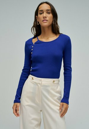 Mujer con una camiseta azul de manga larga acanalada, con botones en el hombro, y pantalones crema de tiro alto con botones dorados.