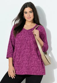 Tunique à motifs floraux violets avec un col en V, des manches trois-quarts et des détails plissés. Accompagnée d'un sac bandoulière beige clair.