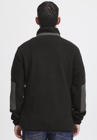 Schwarze Fleecejacke mit hohem Kragen, strukturiertem Gewebe und grauen Kunstlederakzenten an den Ärmeln. Verfügt über lange Ärmel und einen lockeren Schnitt.