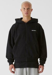 Svart zip-up hoodie i mjukt tyg, med en rymlig huva, framfickor och "LOST YOUTH+"-logotyp i vitt. Grå lager under.