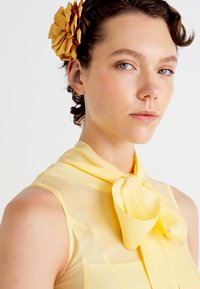 Una giovane donna con i capelli corti e ricci indossa una blusa senza maniche gialla con un grande fiocco e un grosso accessorio per capelli a forma di fiore giallo.