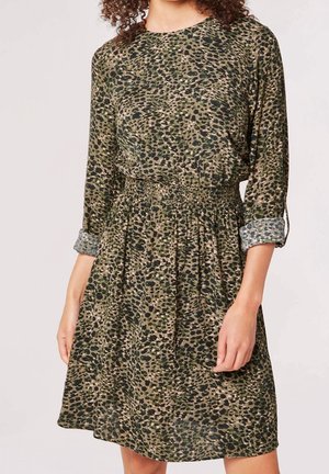 Kvinde iført en grøn og sort leopardprint kjole med trekvartlange ærmer og elastisk talje, stående mod en ensfarvet baggrund.