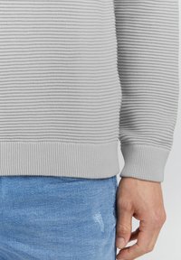 Felpa a coste grigio chiaro con superficie testurizzata, caratterizzata da un orlo e polsini aderenti a coste, abbinata a jeans in denim blu con effetto usurato.