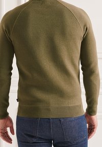 Pull en maille vert olive avec manches raglan et ourlet et poignets côtelés, présenté de dos, associé à un jean en denim bleu foncé.