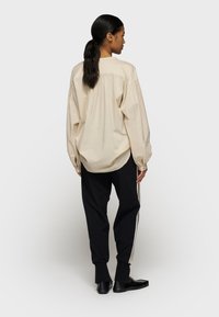 Beige katoenen shirt met lange mouwen, ontspannen pasvorm, achterkant met plooi-detail, gecombineerd met zwarte broek met zijstrepen.