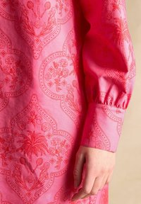 Rotes Kleid mit floralen und Palmenblattmotiven. Der Stoff hat eine glatte Textur und verfügt über schicke Puffärmel sowie ein detailliertes Manschettendesign.