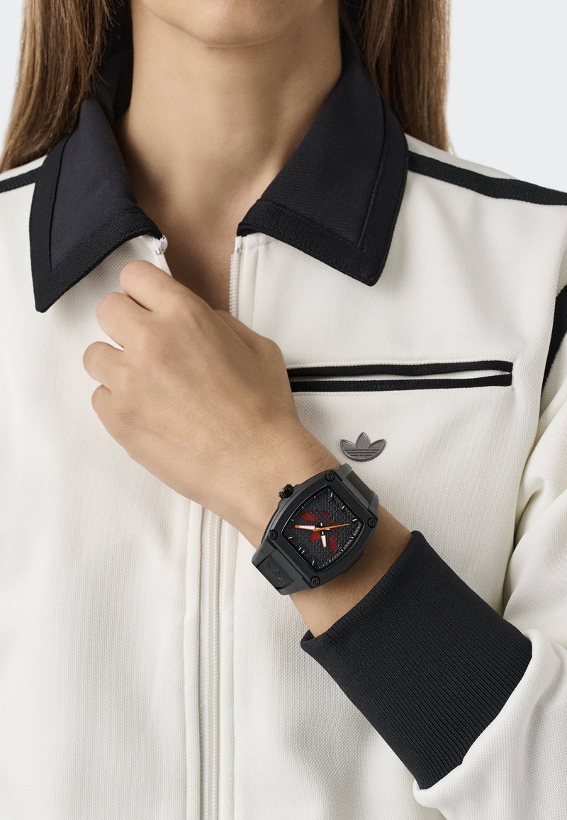 Orologio quadrato nero con dettagli rossi, quadrante testurizzato e hardware in metallo, indossato al polso. Il portatore indossa una giacca bianca con zip e dettagli neri.