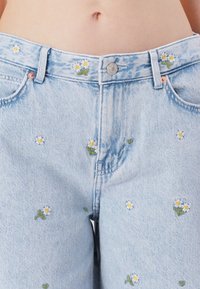 Jean en denim bleu clair avec des marguerites blanches brodées et des feuilles vertes. Comprend une fermeture à bouton et un design classique à cinq poches.
