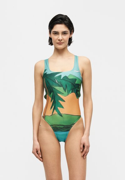 Jeune femme aux cheveux courts et foncés porte un maillot de bain une pièce imprimé de feuilles de palmier vertes et d'un paysage orange et bleu.